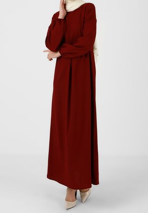 Frau trägt ein langärmeliges, knöchellanges burgunderrotes Kleid mit Puffärmeln und beigefarbenen spitzen Schuhen, steht vor einem schlichten Hintergrund.