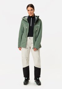 Frau trägt eine grüne Kapuzenjacke, ein schwarzes Reißverschluss-Oberteil, beige-schwarze Skihosen und schwarze Skischuhe und steht vor weißem Hintergrund.