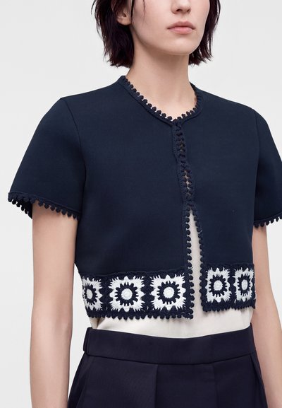 Γυναίκα που φοράει cropped cardigan σε ναυτικό μπλε με κοντά μανίκια και λευκή κεντητή ανθοδέσμη κατά μήκος του τελειώματος, του γιακά και των μανικιών.
