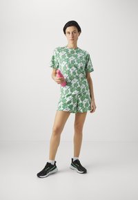 Top de manga corta verde y blanca con estampado de hojas y pantalones cortos a juego. La modelo sostiene una botella de agua rosa. Zapatillas negras con detalles verdes.