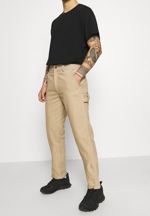 Pantaloni cargo beige con tasche laterali, design a gamba dritta e una texture liscia, abbinati a una t-shirt nera a maniche corte e sneakers nere.