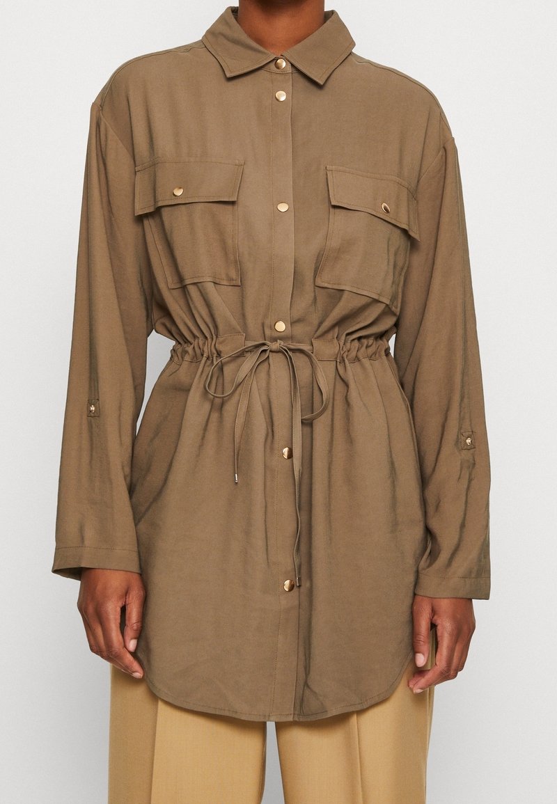 Hnědá košile s dlouhým rukávem, zapínáním na knofy, dvěma kapsami na prsou a elastickým pasem. Měkký materiál s hladkou texturou a zlatými akcenty.