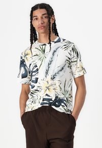 Camisa de manga corta con una base blanca y patrones coloridos de flores y hojas tropicales, hecha de tela ligera, combinada con pantalones marrones acanalados.