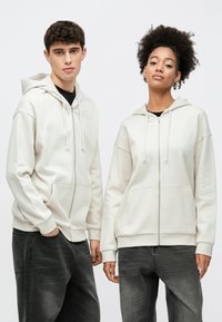 Due persone indossano felpe con cappuccio color crema con cerniera e tasche frontali, abbinate a jeans grigio scuro. Le felpe hanno coulisse e vestibilità morbide.