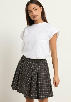 Donna con una maglietta bianca a maniche corte e una gonna plissettata a quadri neri e bianchi, in piedi davanti a uno sfondo chiaro uniforme.