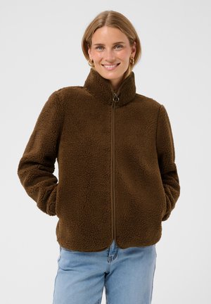 Femme souriante portant une veste polaire marron avec fermeture éclair devant et un jean bleu clair, mains dans les poches de la veste, arrière-plan neutre.