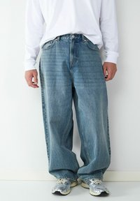 Weit geschnittene blaue Jeans aus Denim mit ausgewaschenem Finish, Fünf-Taschen-Design und Metallknopfverschluss, kombiniert mit einem langärmligen weißen Hemd und Turnschuhen.