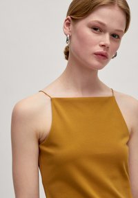 Mosterdgele mouwloze top met een rechte halslijn en dunne bandjes, met een zachte, gladde textuur. Geen patronen of versieringen.