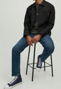 Svart knapplös jacka med ficka fram, grön skjorta under, blå raka jeans och marinblå högstövlar på en pall.