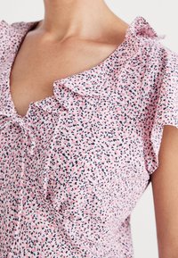 Rosa Bluse mit gebauschter Halslinie und kurzen Ärmeln. Der Stoff zeigt kleine schwarze und rote Blumenmuster auf einem hellen Hintergrund.