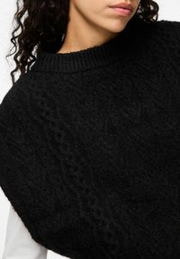 Svart stickad tröja med en texturerad design. Har en hög, ribbad halsringning och en lös passform, vilket visar ett tjockt, mysigt material.