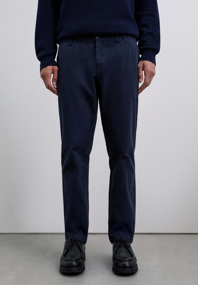 Pantalones de algodón azul marino con pierna recta, con bolsillos laterales y cierre de botón, combinados con zapatos negros de cordones.