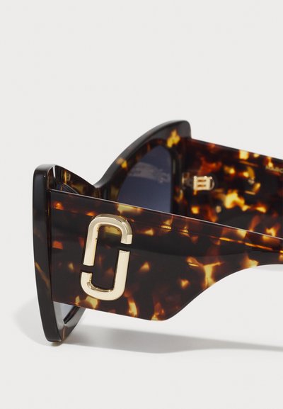 Sonnenbrille im Schildpatt-Design mit einem kontrastierenden goldenen Logo-Akzent auf dem Rahmen, ausgestattet mit einer glänzenden Oberfläche und getönten Gläsern.