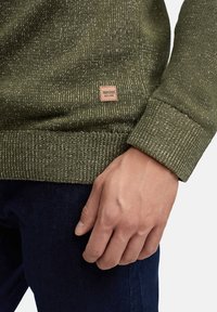 Olivengroene gebreide trui met textuurstof, voorzien van een geribde zoom en manchetten, en een klein bruin lederen label met de tekst "INVI CODE."