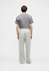 Άνδρας που φορά γκρι μπλουζάκι και ανοιχτό γκρι φόρμα, στέκεται με την πλάτη στην κάμερα μπροστά σε απλό λευκό φόντο.