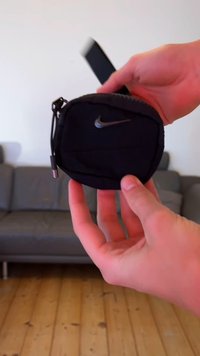 Μαύρη υφασμάτινη θήκη με φερμουάρ, με το ασημί λογότυπο της Nike. Ο σχεδιασμός είναι στρογγυλός και διαθέτει λουράκι για τον καρπό, με ομαλή υφή.