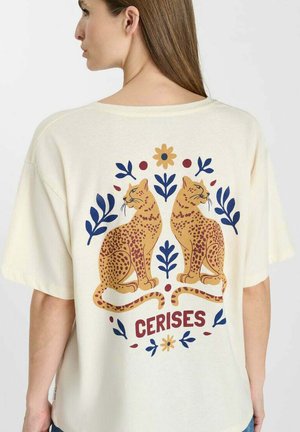 Vrouw draagt een crèmekleurig t-shirt met twee zittende luipaarden die wegkijken, omringd door blauw- en geelgekleurde bloemmotieven en het woord "CERISES."