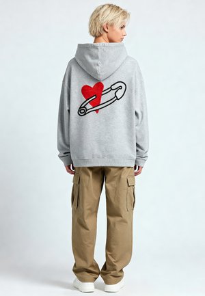 Grijze hoodie met een rode hart- en veiligheidsspeldafbeelding op de rug, gecombineerd met khaki cargobroek en witte sneakers.
