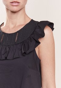 Černý top bez rukávů s texturovaným volánovým výstřihem a zdobením na ramenou, vyrobený z lehké látky s hladkým povrchem.