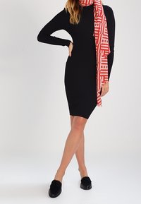 Zwarte geribbelde jurk met lange mouwen, gecombineerd met een rood-wit patroon sjaal. Model draagt zwarte instapschoenen met bontaccenten.