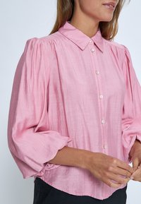 Blusa rosa con colletto, maniche a palloncino, tessuto testurizzato e chiusura a bottoni. Presenta un orlo curvo e una leggera lucentezza, evidenziando la morbidezza del materiale.