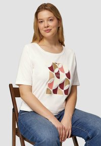 Witte katoenen t-shirt met korte mouwen met een geometrische print in bordeaux, roze, goud en beige. Model zittend op een houten stoel.