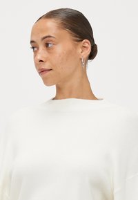 Weißer gerippter Pullover mit hohem Kragen und überschnittenen Schultern, kombiniert mit silbernen Creolen. Die strukturierte Stoffqualität sorgt für visuelle Auflockerung.