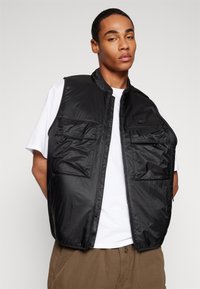 Μαύρο νάιλον γιλέκο με δύο μπροστινές τσέπες, κλείσιμο με φερμουάρ και διακριτική λεπτομέρεια λογότυπου, φορεμένο πάνω από ένα άσπρο μπλουζάκι, με ελαφρύ, υφασμάτινο σχέδιο.