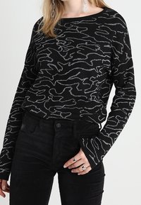  Schwarzer Pullover mit silbernem abstraktem Wellenmuster, langen Ärmeln und weitem Ausschnitt. Kombiniert mit schwarzen Cordhosen, akzentuiert mit Ringen.