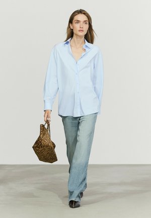 Femme marchant portant une chemise bleu clair boutonnée, un jean bleu délavé, des chaussures noires et tenant un sac à main imprimé léopard dans sa main gauche.