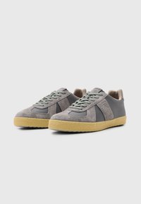 Sneakers grigi in suede con un design testurizzato, dettagli verdi e una suola in gomma marrone chiaro. Presentano lacci piatti e un colletto imbottito.