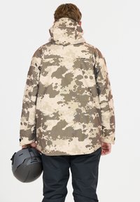 Kamikaze takki vaale ja ruske pakke, jeb de splattered patterns, hood, ja zipp jaas taap. Kaali pants aur helmet haath mein pakre hue.