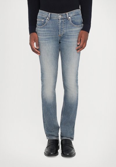 Világoskék farmernadrág, slim fit szabású, öt zsebbel, enyhe kopással a combokon, fémből készült kiegészítőkkel. Fekete cipővel viselve.