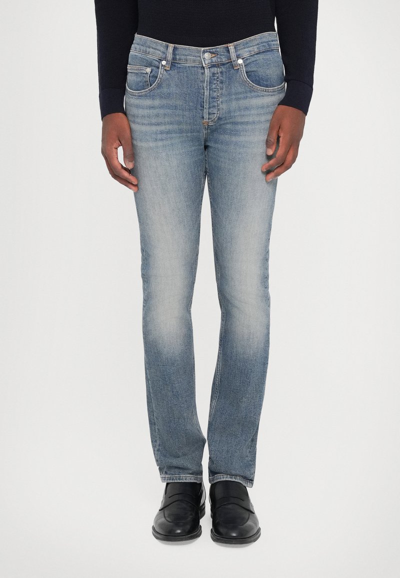 Világoskék farmernadrág, slim fit szabású, öt zsebbel, enyhe kopással a combokon, fémből készült kiegészítőkkel. Fekete cipővel viselve.