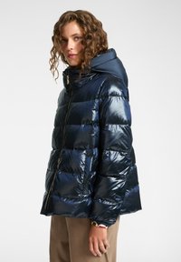 Chaqueta acolchada marina con una superficie brillante y acolchada, un cuello alto y cierre frontal con cremallera. Cuenta con capucha y bolsillos laterales.