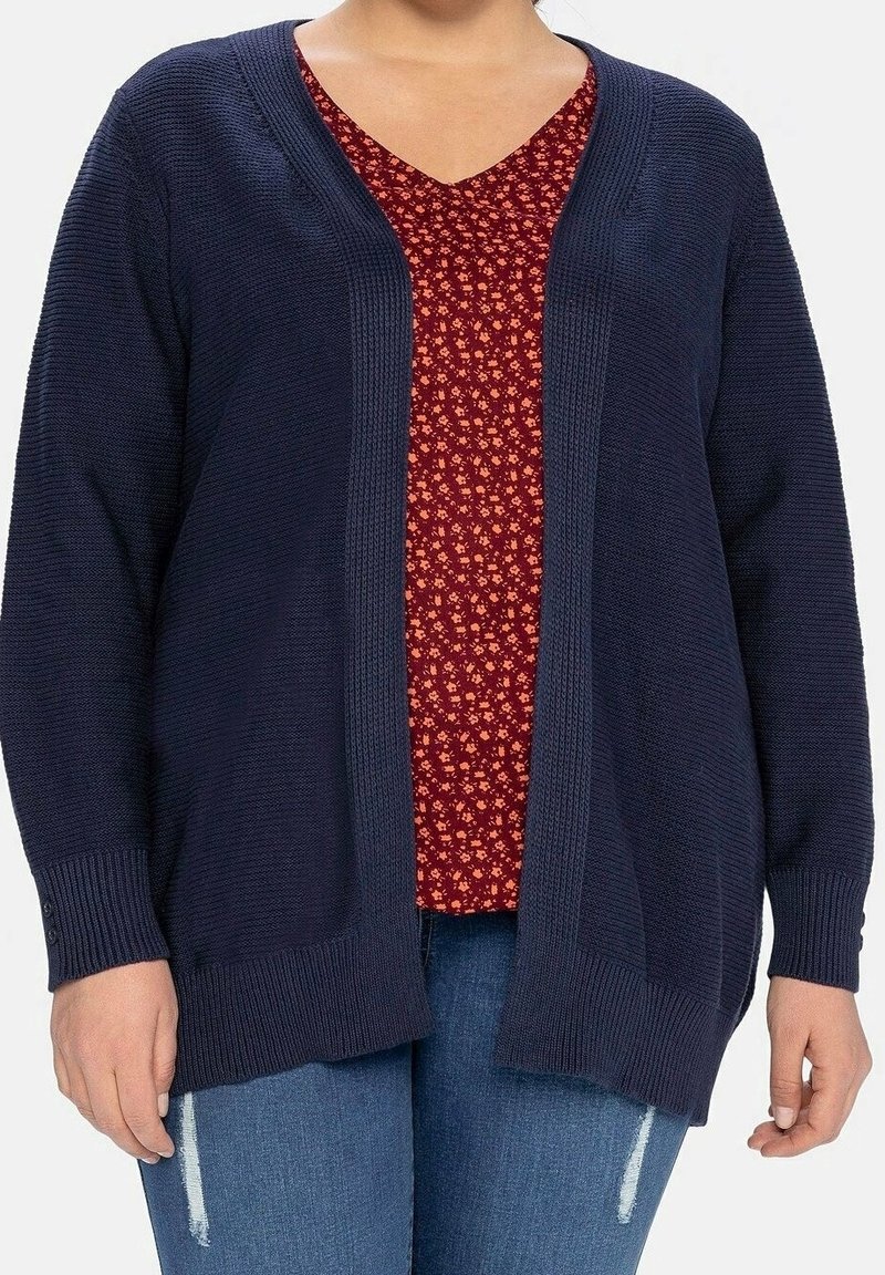Femme portant un cardigan bleu marine ouvert sur un top rouge à motifs floraux et col en V avec un jean bleu délavé.