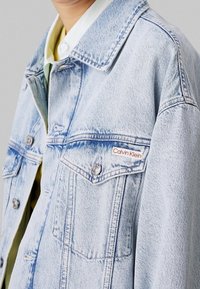 Ljusblå denimjacka med två bröstfickor, silverfärgad hårdvara och en märkesskylt på vänster ficka. Slät yta och avslappnad passform.