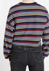 Person som bär en långärmad skjorta med horisontella ränder i marinblått, rött, gult och vitt, tillsammans med svarta denimjeans, sedd bakifrån.