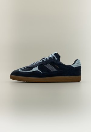 Μαύρο και γκρι sneaker από σουέντ με χαμηλό προφίλ, λαστιχένια σόλα, πλαϊνές ρίγες και κορδόνια, εμφανιζόμενο από την εξωτερική πλευρά σε ουδέτερο φόντο.