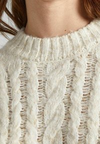 Kerrosneuletakki kermanvalkoisena; siinä on teksturoidut kuviot, resorikaulus ja pehmeä, pörröinen materiaali.