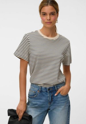 Jeune femme aux cheveux châtain clair porte un t-shirt rayé beige et noir et un jean bleu, tenant une pochette noire pliée dans une main.