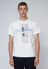 Weißes Baumwoll-T-Shirt mit einem zentralen Grafikmotiv von Palmen und dem Text "LES HOMMES" in Blau. Kurze Ärmel und runder Halsausschnitt.