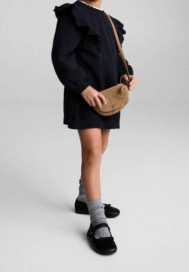 Enfant portant une robe noire à épaules volantées, des chaussettes grises, des chaussures Mary Jane noires et portant un petit sac marron en bandoulière.