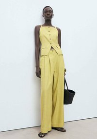 Completo composto da gilet a righe gialle e pantaloni a gamba larga con bottoni e tasche discrete. Modella che tiene una borsa intrecciata nera, in piedi contro uno sfondo neutro.