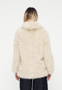Cappotto in pelliccia ecologica beige, con una texture morbida e soffice, colletto ampio e spalle scese. L'orlo è più lungo sul retro per una maggiore copertura.