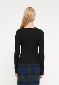 Γυναίκα με μακριά ξανθά μαλλιά, φοράει κολλητό μαύρο μακρυμάνικο τοπ και μπλε-πράσινη καρό φούστα, γυρισμένη πλάτη σε λευκό φόντο.