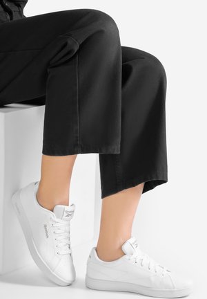 Weiße Ledersneaker mit glatter Textur, ausgestattet mit einer Schnürung und dem Reebok-Logo an der Seite und auf der Zunge, kombiniert mit schwarzen Hosen.