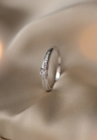 Bague en argent avec un diamant rond central et deux rangées de petits diamants le long de l'anneau, reposant sur un fond de tissu beige doux.