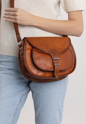 Vrouw draagt lichtblauwe spijkerbroek en beige top met een bruine leren crossbodytas met gespsluiting die op haar heup rust.