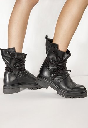 Stivaletti alla caviglia in pelle nera testurizzata con lacci avvolgenti e suole in gomma spessa indossati su gambe nude su sfondo bianco.